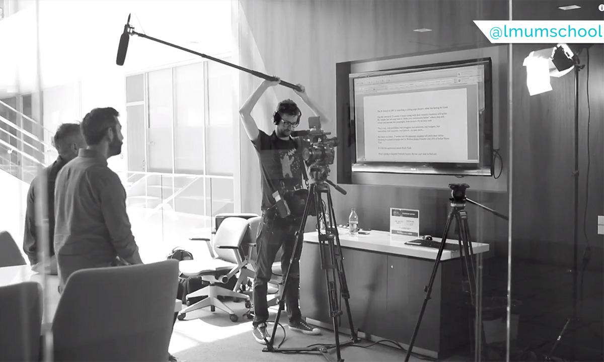 A student holding a boom mic and standing behind a camera while two faculty are being filmed