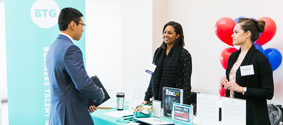 A student in professional attire speaking to representatives behind at a booth at a career fair